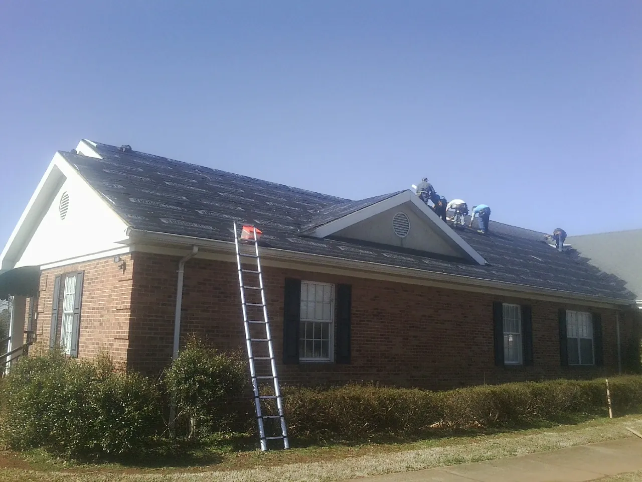 Crew tearing shingles off a brick ranch roof with a ladder set — Metal Roof Repair work by our crew in Cedarbluff